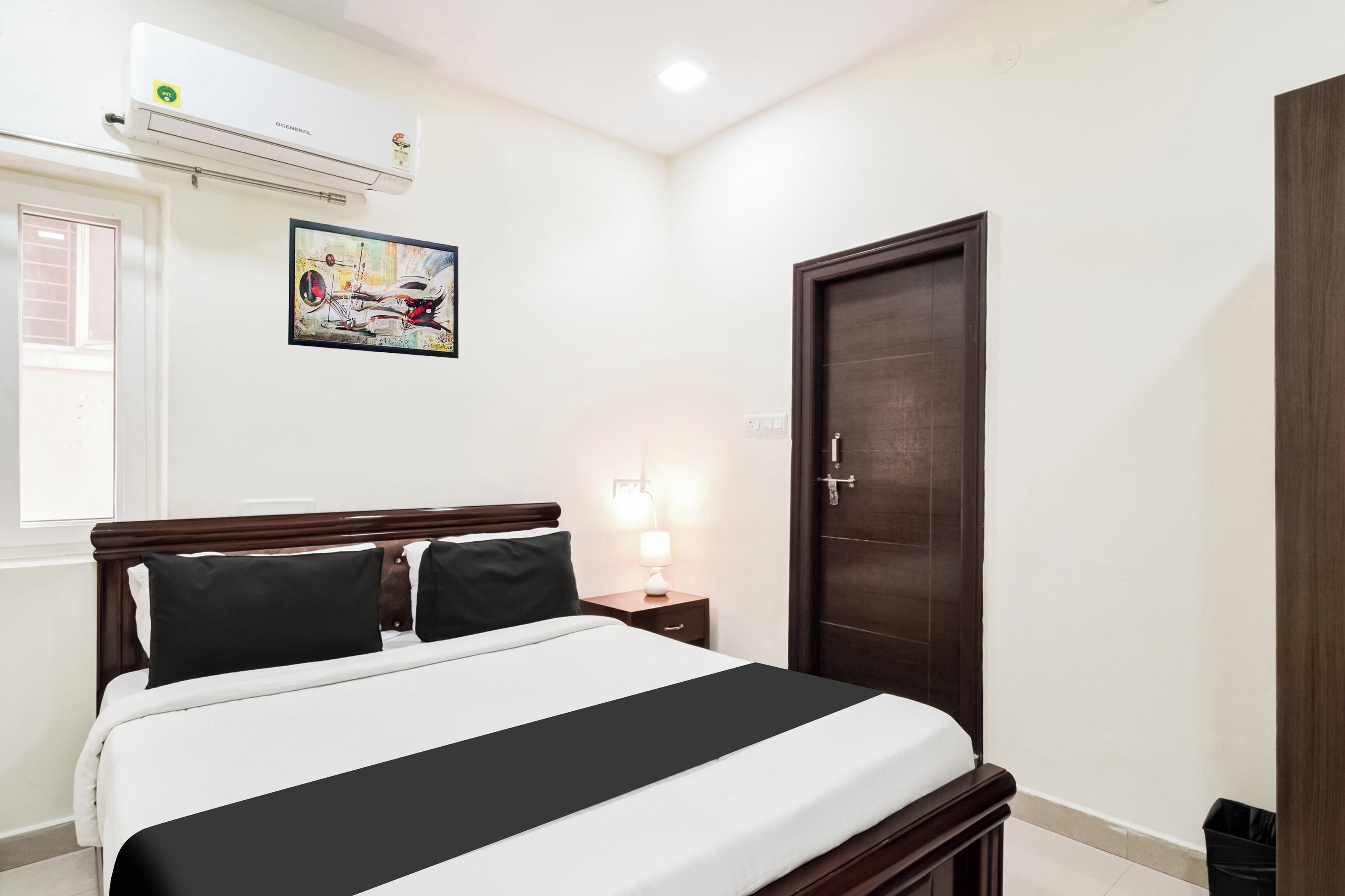 A hotel room with a bed, a nightstand, a lamp, a door, and an air conditioning unit.