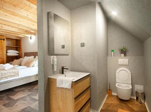 Ein Schlafzimmer mit einem Holzschrank, einem Bett mit weißer Bettwäsche, einer Holzkommode mit Spiegel und einem Badezimmer mit Toilette und Waschbecken.