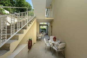 A narrow outdoor corridor with a staircase leading to a balcony, a dining area with tables and chairs, and a potted plant. - Apartments Botica- Double Room (Zupa dubrovacka)