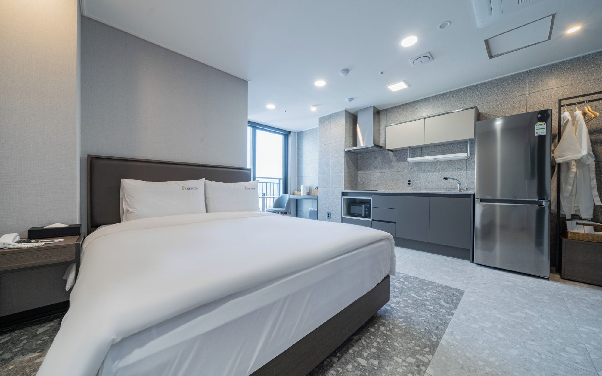 Una habitación de hotel moderna con una cama grande, un escritorio con teléfono, una kitchenette con refrigerador y una ventana con vistas.