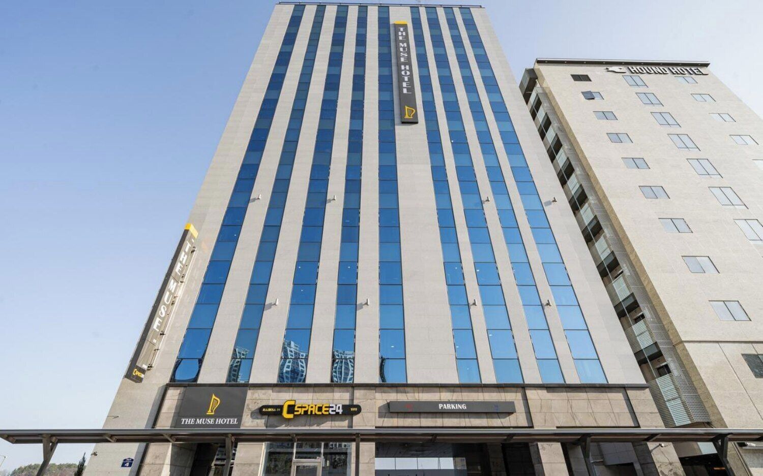 A tall hotel building with a modern design, featuring a prominent sign for The Muse Hotel and a visible parking area.