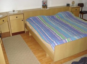 A single bed with a striped bedspread, a bedside table with a radio, and a chair in a room with wooden flooring and a plain wall. - Apartmani Cihorich - One Bedroom Apartment (Dugi Rat)