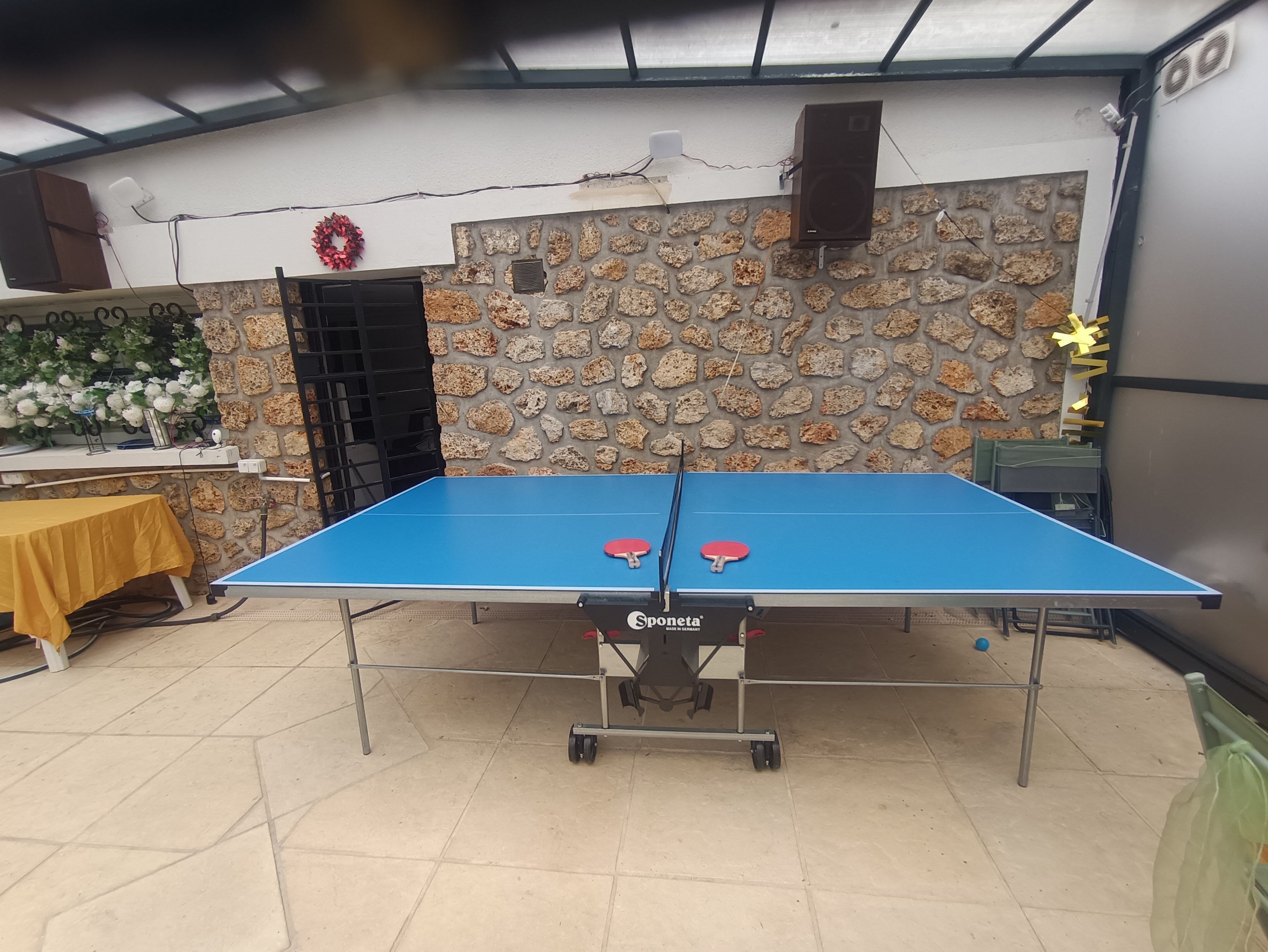 A ping pong table with two paddles, a stone wall, and a speaker mounted on the wall.