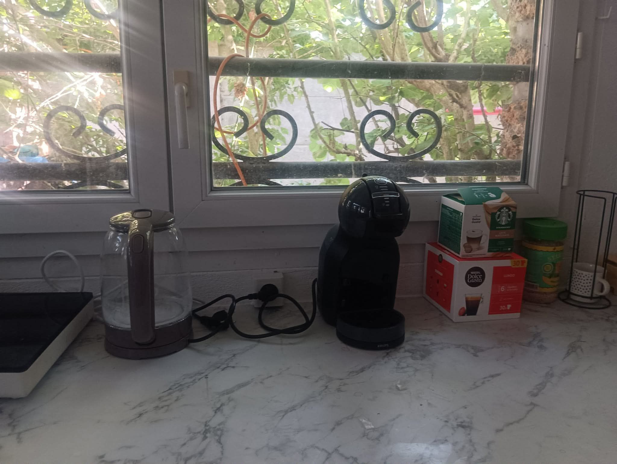 Un plan de travail de cuisine avec une machine à café, un récipient en verre et une boîte de capsules de café Dolce Gusto.