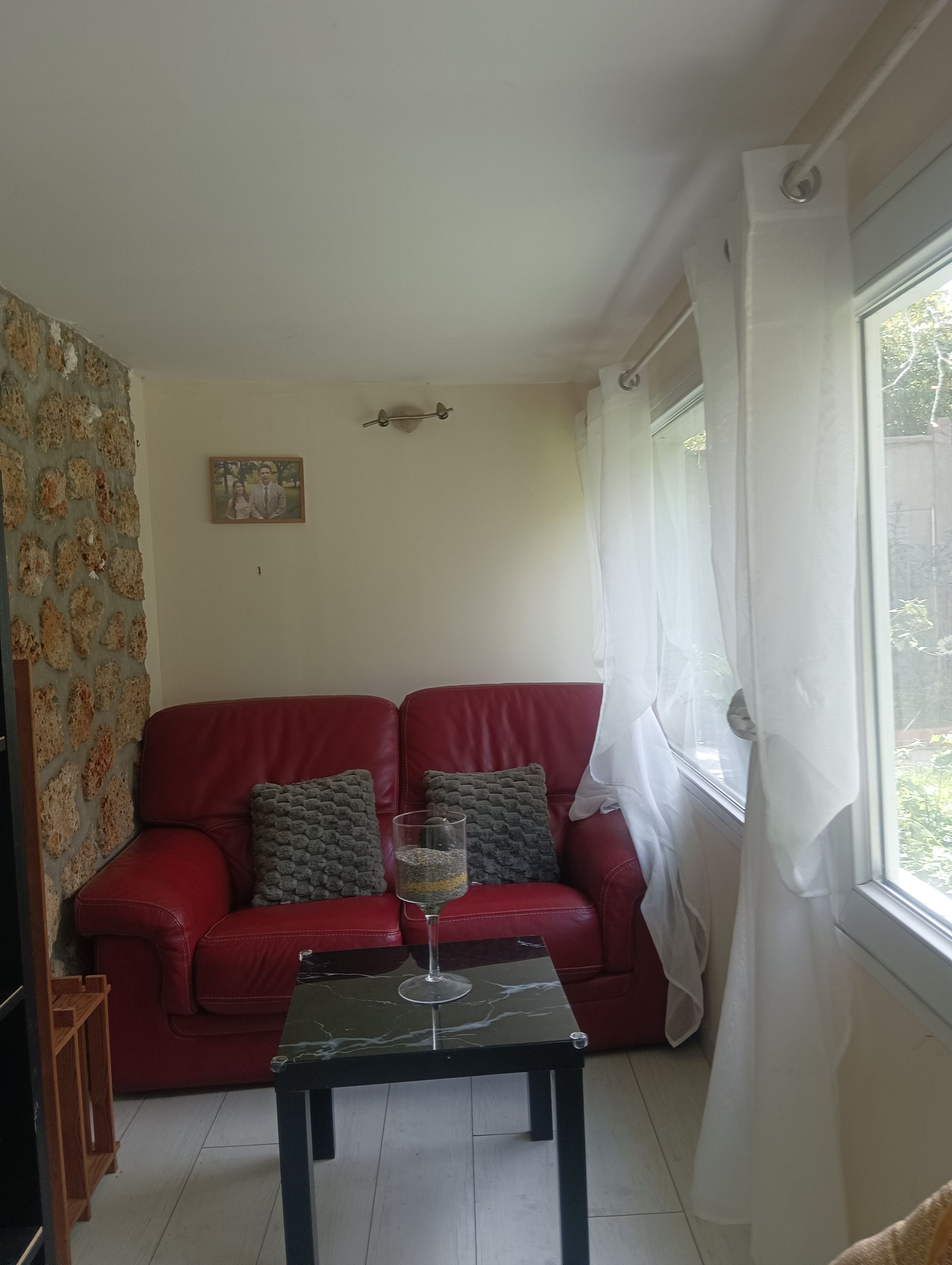 Un sofá de cuero rojo con cojines, una mesa de vidrio con una copa de vino y una ventana con cortinas blancas.