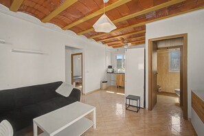 A modern living room with a black sofa, white coffee table, and a pendant light. There is a kitchen area visible through an open door.