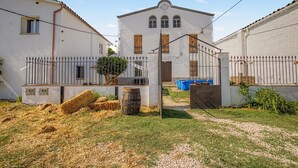 Un edificio blanco con una entrada central, un patio cercado con fardos de heno y un contenedor de basura azul.