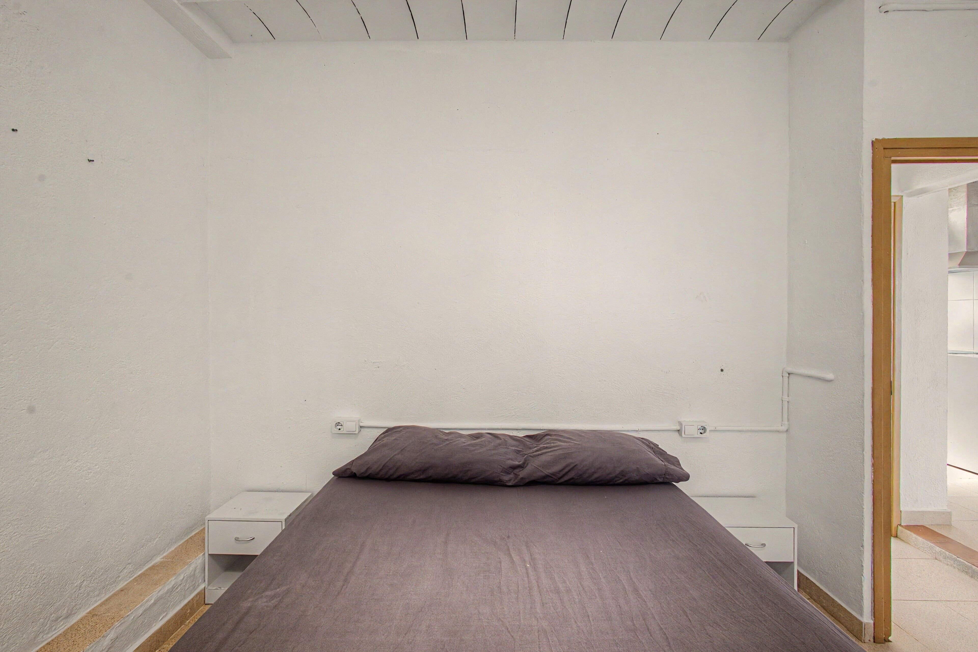 A small, single-bed room with a plain white wall, a wooden door frame, and a bedside table with a drawer.