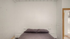 Una habitación pequeña con una cama individual, una pared blanca sencilla, un marco de puerta de madera y una mesita de noche con cajón.