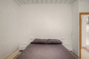 A small, single-bed room with a plain white wall, a wooden door frame, and a bedside table with a drawer. - Rural house 'Maset De La Salut' with mountain views, Wi-Fi, and air conditioning (Piera)