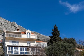A two-story building with balconies, a mountain backdrop, and a clear sky. - Apartment M&K - Two Bedroom Apartment With Balcony and Sea View (Zupa dubrovacka)