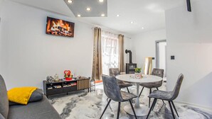 Een moderne woonkamer met een open haard, een ronde eettafel met stoelen, een bank met een geel kussen en een televisie aan de wand.