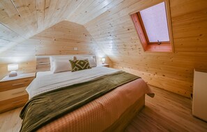 Ein Schlafzimmer mit holzverkleideter Schräge, einem Bett mit weißer Bettwäsche und einem grünen Kissen, einem Nachttisch mit Lampe und einem Fenster mit rotem Rahmen.
