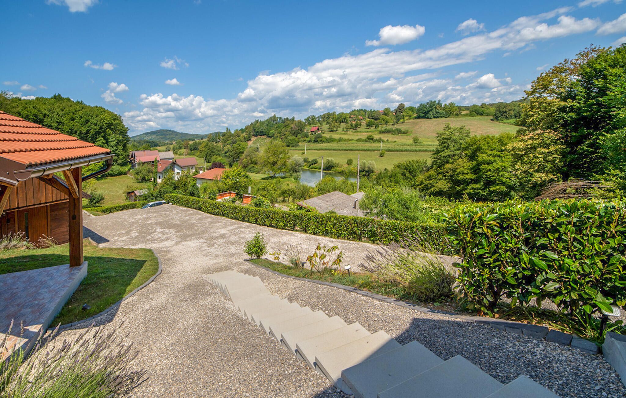 A rural landscape with a gravel driveway leading to a wooden building, surrounded by lush greenery and a clear sky.