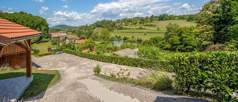 A rural landscape with a gravel driveway leading to a wooden building, surrounded by lush greenery and a clear sky.