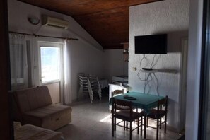 A small, compact room with a wooden ceiling, a sofa, a dining table with chairs, a television mounted on the wall, and a window with curtains. - Apartments Roza - Studio Apartment with Sea View (Herceg Novi)