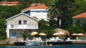 A coastal building with a stone facade, a balcony, and a thatched-roof structure with lounge chairs and umbrellas. - Apartments Roza - Standard Double Room with Shared Bathroom (Herceg Novi)
