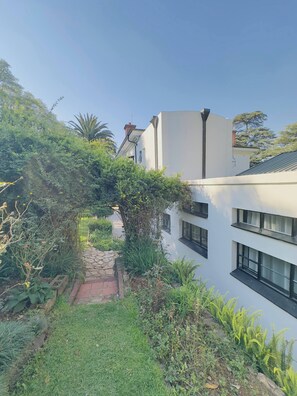 Un edificio moderno con fachada blanca, múltiples ventanas y un sendero que lleva a un jardín con diversas plantas y un pequeño árbol.