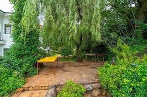 Un patio de ladrillo con una mesa y sillas amarillas, rodeado de abundante follaje y árboles.
