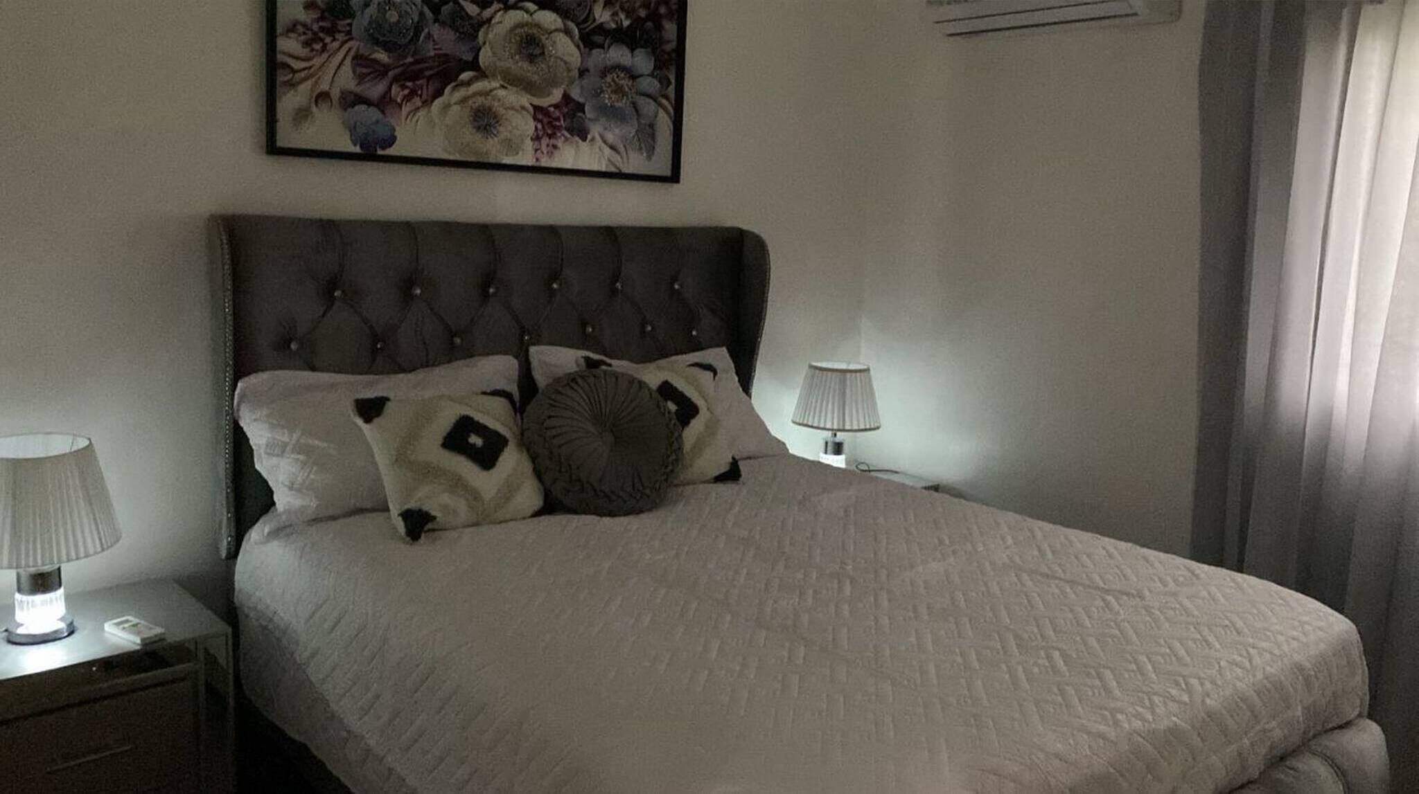 Una cama bien hecha con un cabecero capitonado, dos lámparas de noche y una obra de arte floral en la pared.