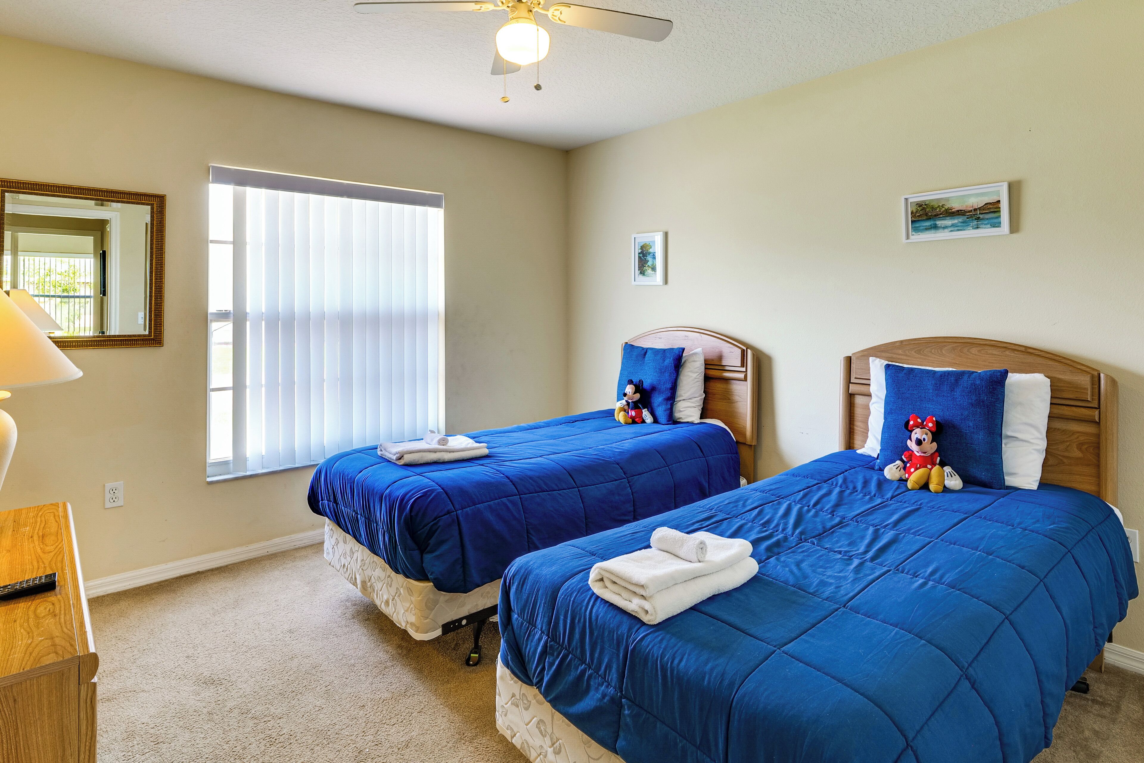 A hotel room with two beds, a ceiling fan, a window with blinds, and a small desk with a lamp.