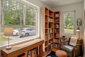 Ein Zimmer mit großem Fenster, einem Holztisch, einem Bücherregal, einem Sessel und einer Wanduhr.