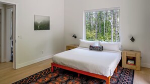 Un dormitorio con cama, ventana con vista a árboles, mesita de noche con lámpara y alfombra.