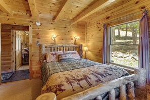 A wooden cabin bedroom with a bed, two wall-mounted lamps, a window with curtains, and a door leading to another room. - Incredible Views! Log Cabin w/ Wraparound Deck (Angel Fire)