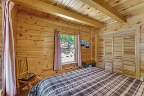 Ein Schlafzimmer einer Holzhütte mit einem Bett, einem Fenster mit Vorhängen, einem kleinen Tisch und einem Kleiderschrank.