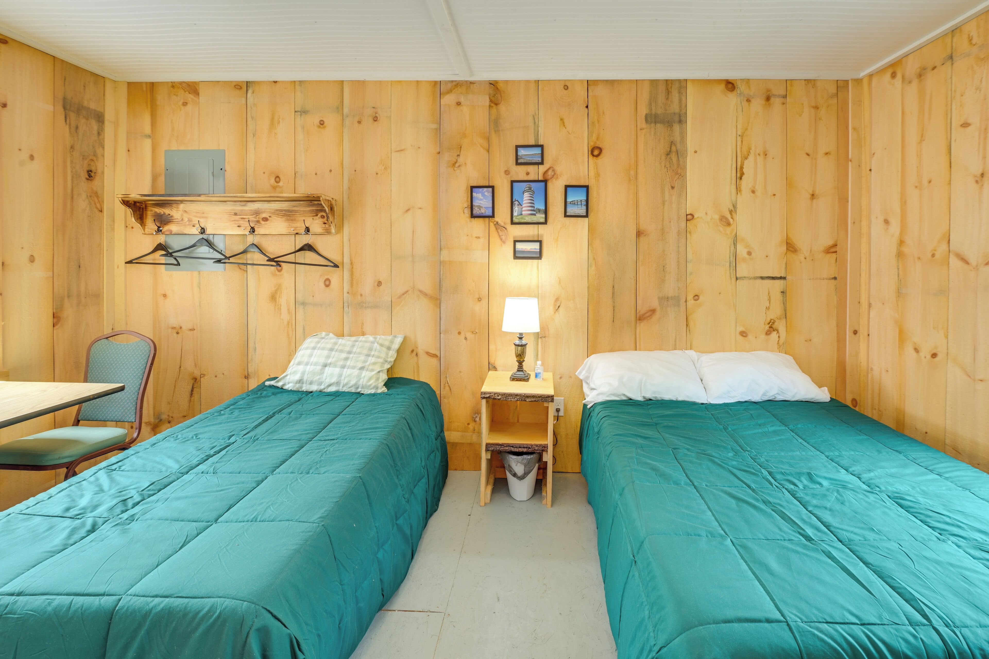 A room with two beds, a table, a chair, a lamp, and a small trash bin.