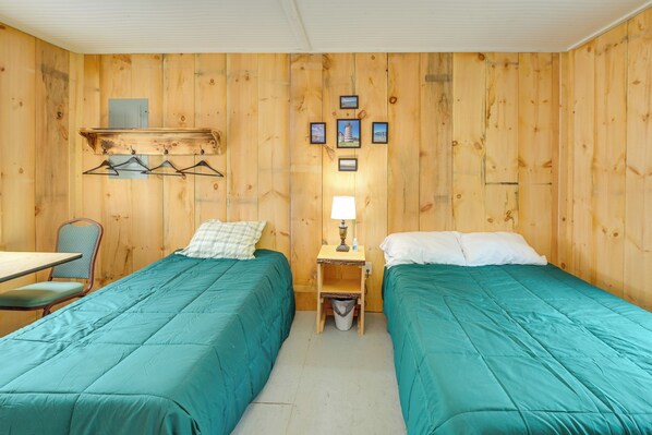 A room with two beds, a table, a chair, a lamp, and a small trash bin.