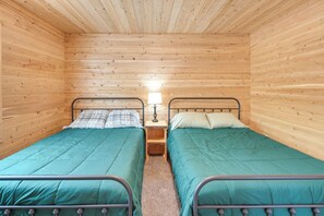 A small, cozy room with two single beds, each with a metal frame and a green bedspread. There is a nightstand with a lamp between the beds.