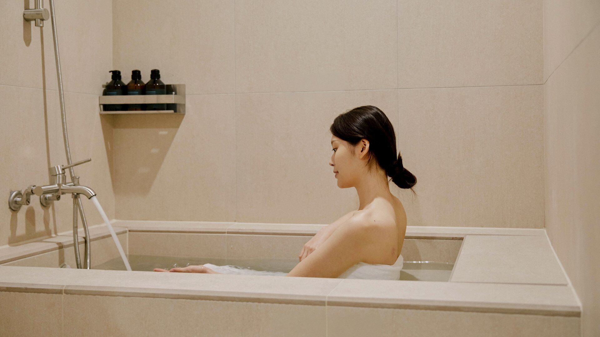 A person in a bathtub with a showerhead and a shelf with bottles.