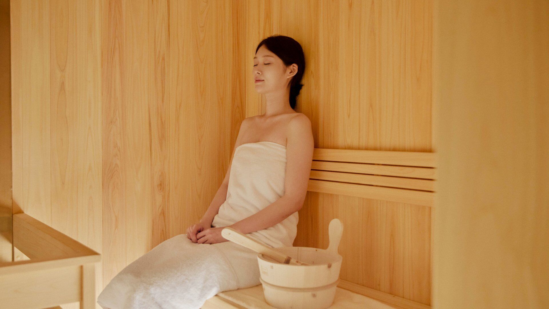 A person seated in a wooden sauna with a wooden bench and a bowl.