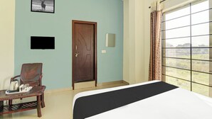 A hotel room with a bed, a chair, a table with a kettle, a picture on the wall, a door, and a window with curtains.