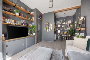 A modern living room with a flat-screen TV, bookshelves, a dining area, and a sofa. - Spacious 1BR in Near Arthur's Seat & Royal Mile (Edinburgh)