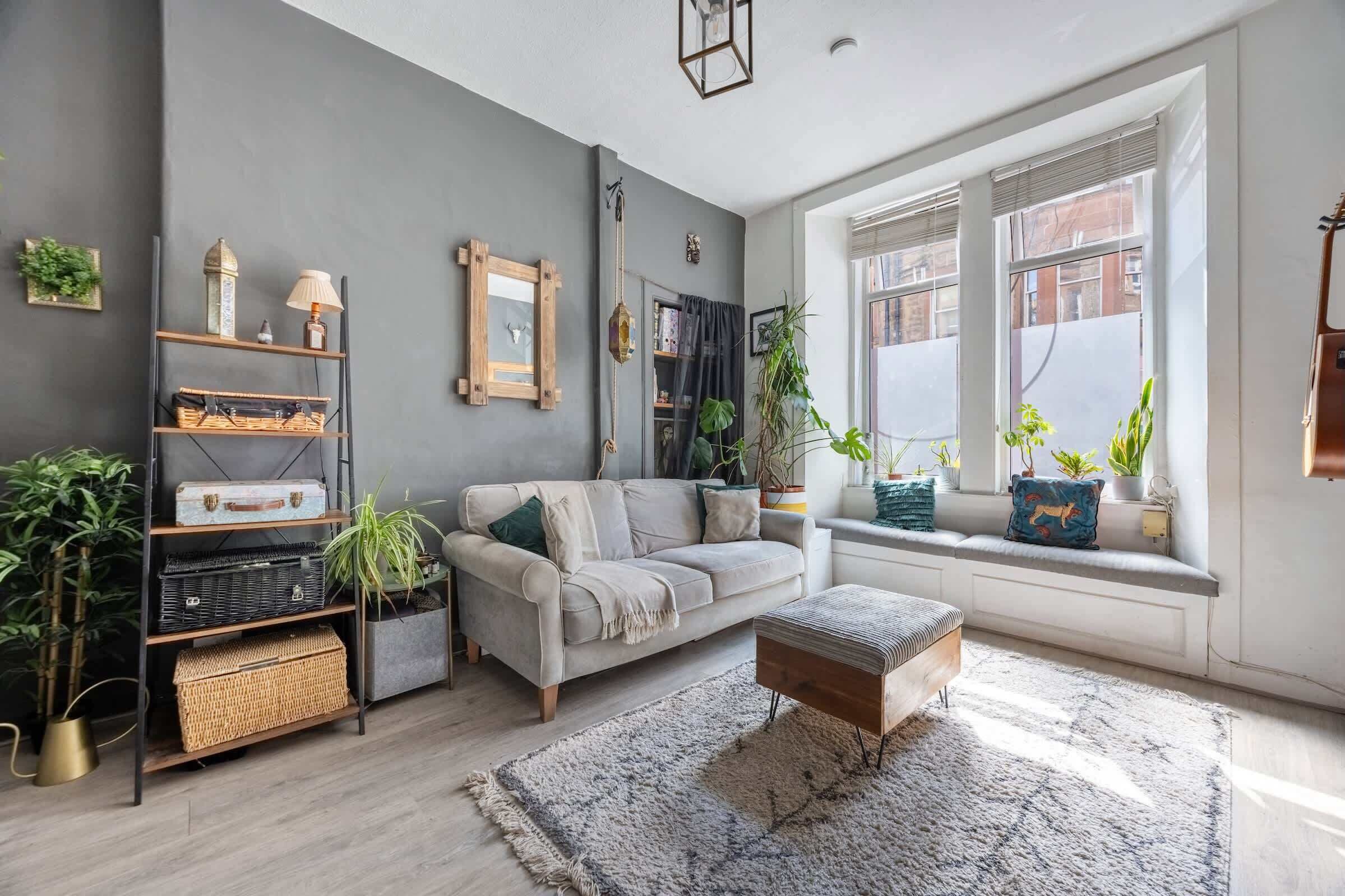 Un salon moderne avec un canapé gris, un banc en bois et une bibliothèque. Des plantes sont posées sur le rebord de la fenêtre, et une grande fenêtre offre une vue sur l’extérieur.