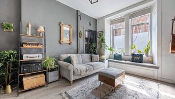 Un salon moderne avec un canapé gris, un banc en bois et une bibliothèque. Des plantes sont posées sur le rebord de la fenêtre, et une grande fenêtre offre une vue sur l’extérieur.