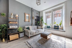 Una sala de estar moderna con un sofá gris, un banco de madera y una estantería. Hay plantas en el alféizar de la ventana y un ventanal que ofrece vistas al exterior.