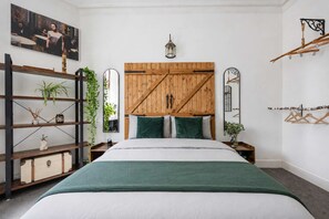 Quarto com cabeceira de madeira, cama com almofadas verdes, mesa de cabeceira com abajur e espelho fixado na parede.