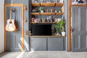 A room with a grey door, a guitar leaning against it, a TV on a wooden shelf, and a potted plant. - Spacious 1BR in Near Arthur's Seat & Royal Mile (Edinburgh)