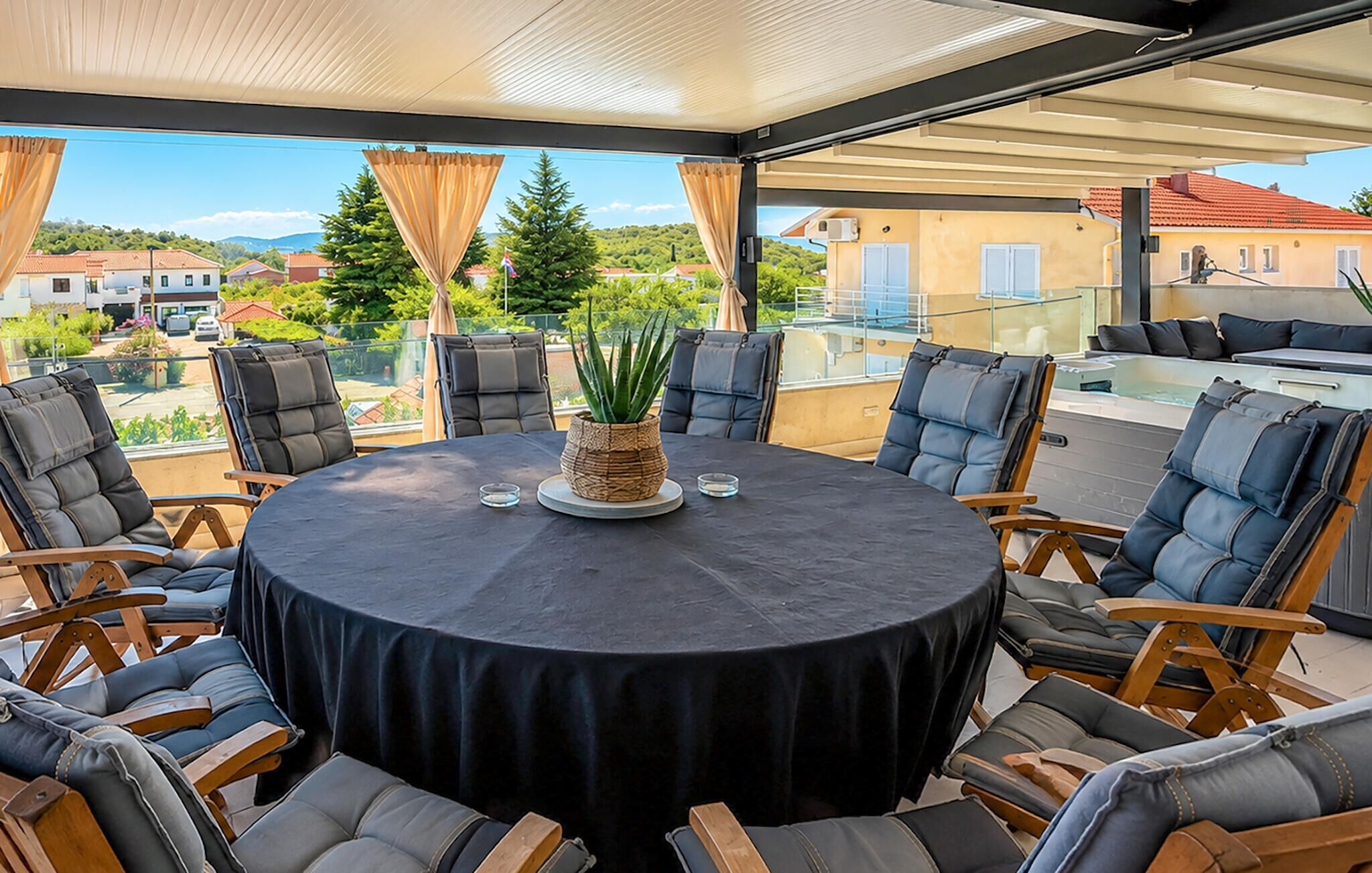Una mesa redonda con mantel negro, rodeada por seis sillas con estructura de madera y cojines grises, ubicada en un balcón con vistas a casas y vegetación.