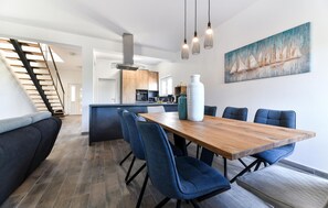 Ein moderner Essbereich mit Holztisch und blau gepolsterten Stühlen, im Hintergrund eine Küche und eine Treppe, die zu einer anderen Ebene führt.
