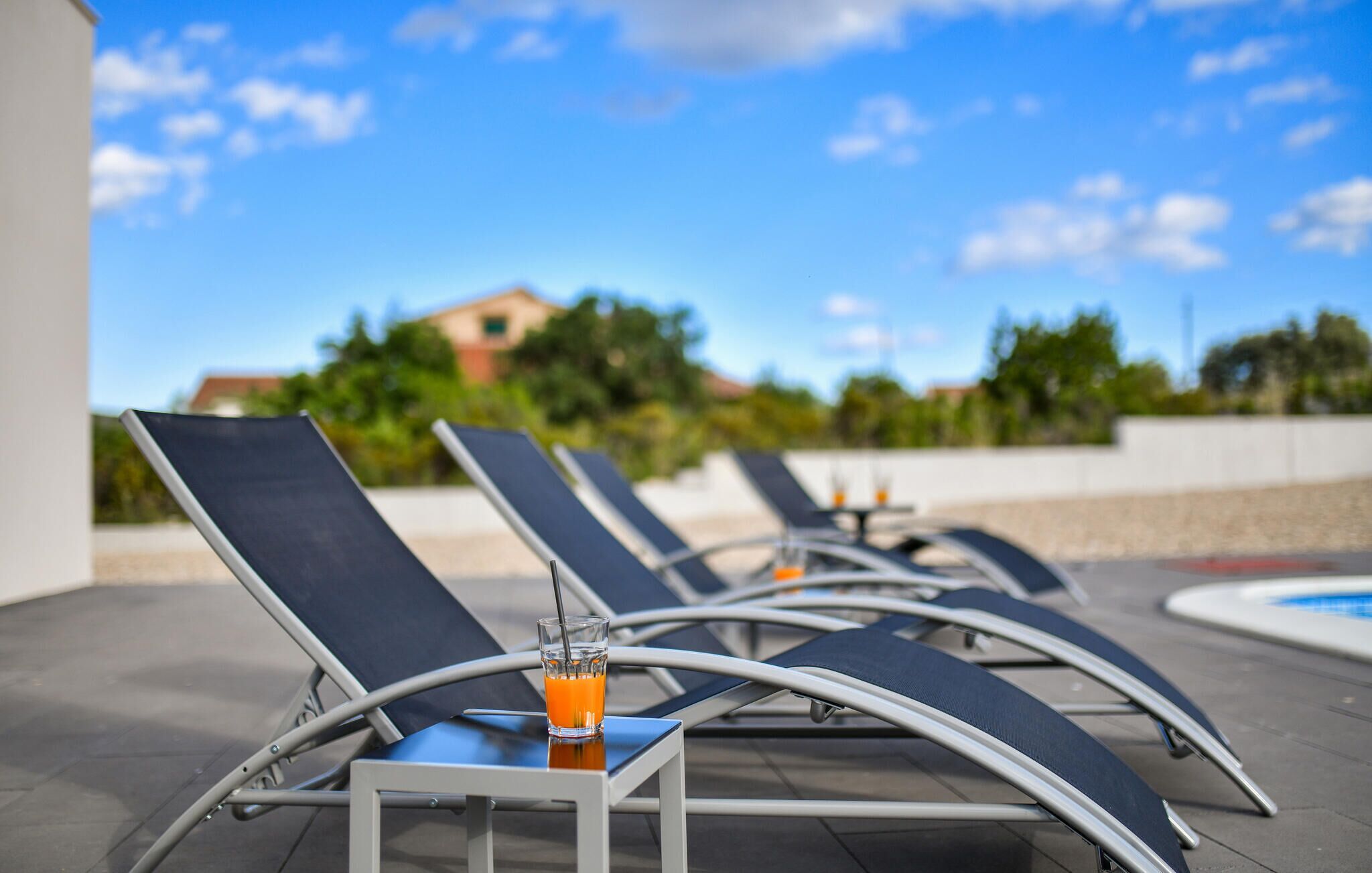 A row of sun loungers with a glass of orange juice on a table, a pool in the background, and a clear sky.