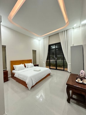 A modern hotel room with a large bed, a wooden headboard, a dining table, and a sliding glass door leading to a balcony.