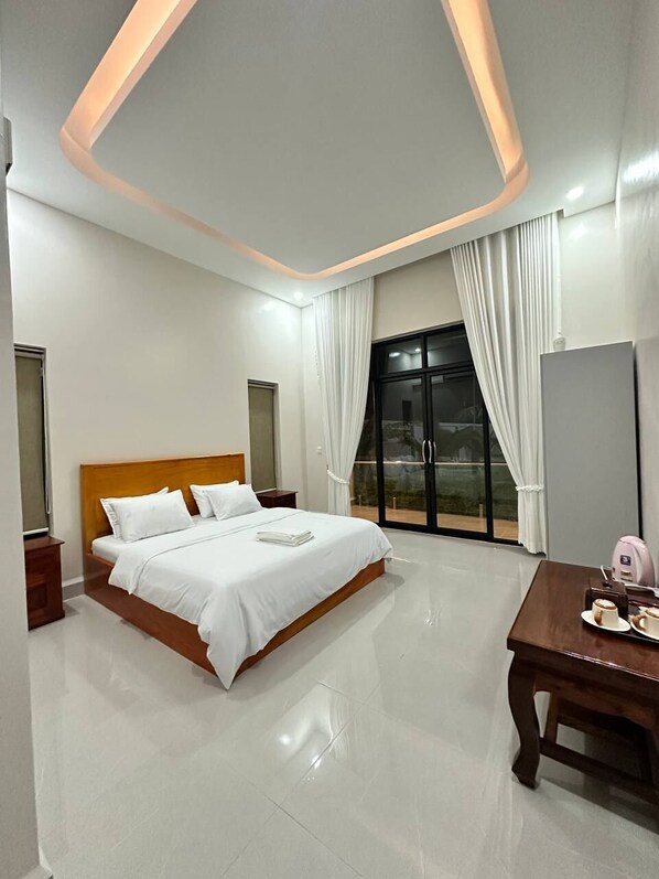 A modern hotel room with a large bed, a wooden headboard, a dining table, and a sliding glass door leading to a balcony.