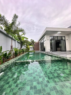 A swimming pool with a tiled floor, surrounded by greenery and a modern building with large windows.