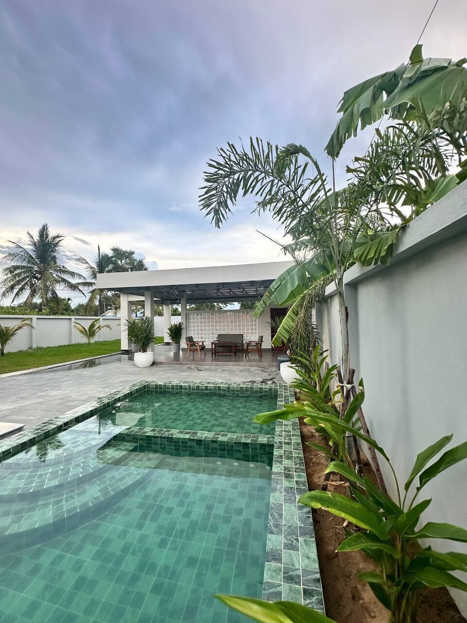 Una piscina con borde de azulejos, rodeada de exuberante vegetación y una zona de estar cubierta.