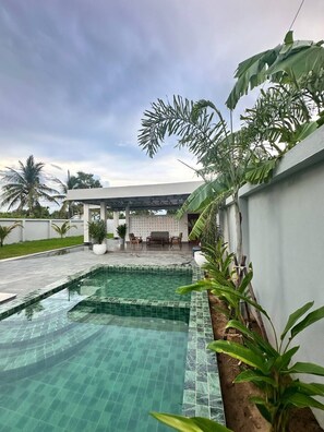 A swimming pool with a tiled edge, surrounded by lush greenery and a covered seating area.