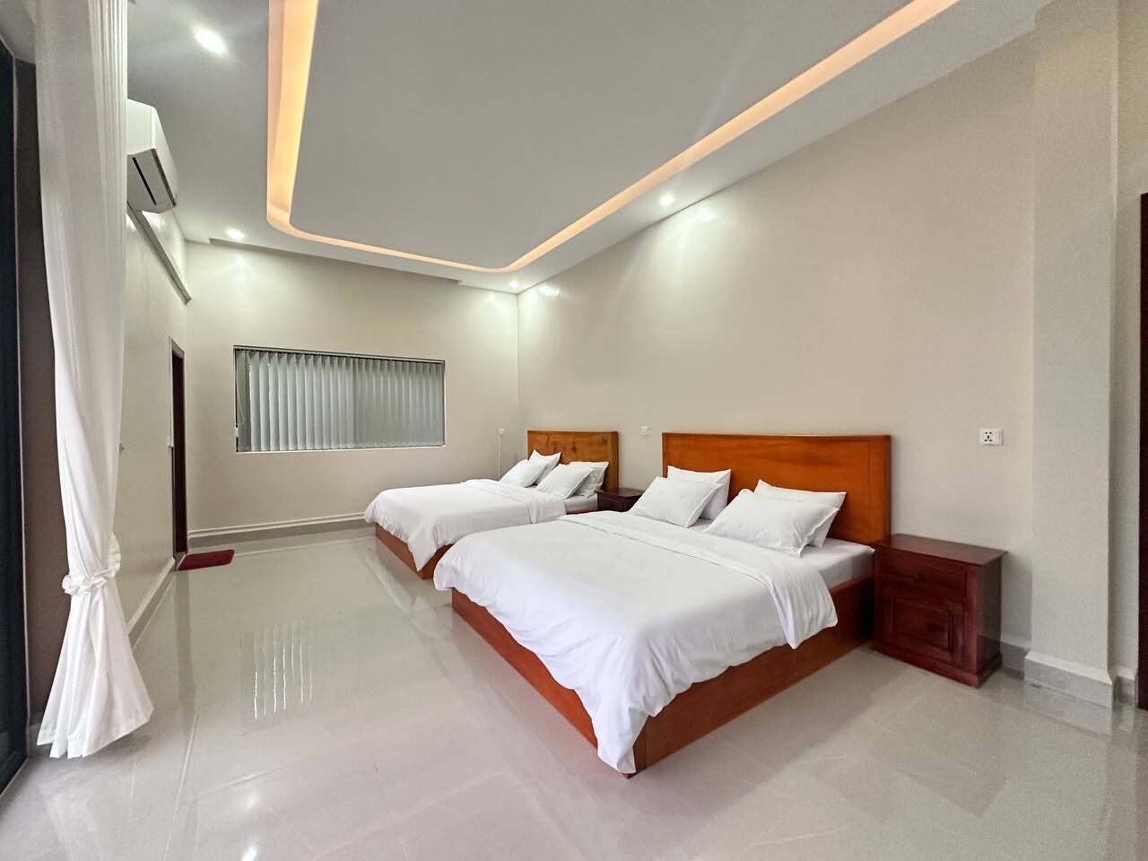 A hotel room with two beds, a large window with blinds, a wooden nightstand, and a modern ceiling design.